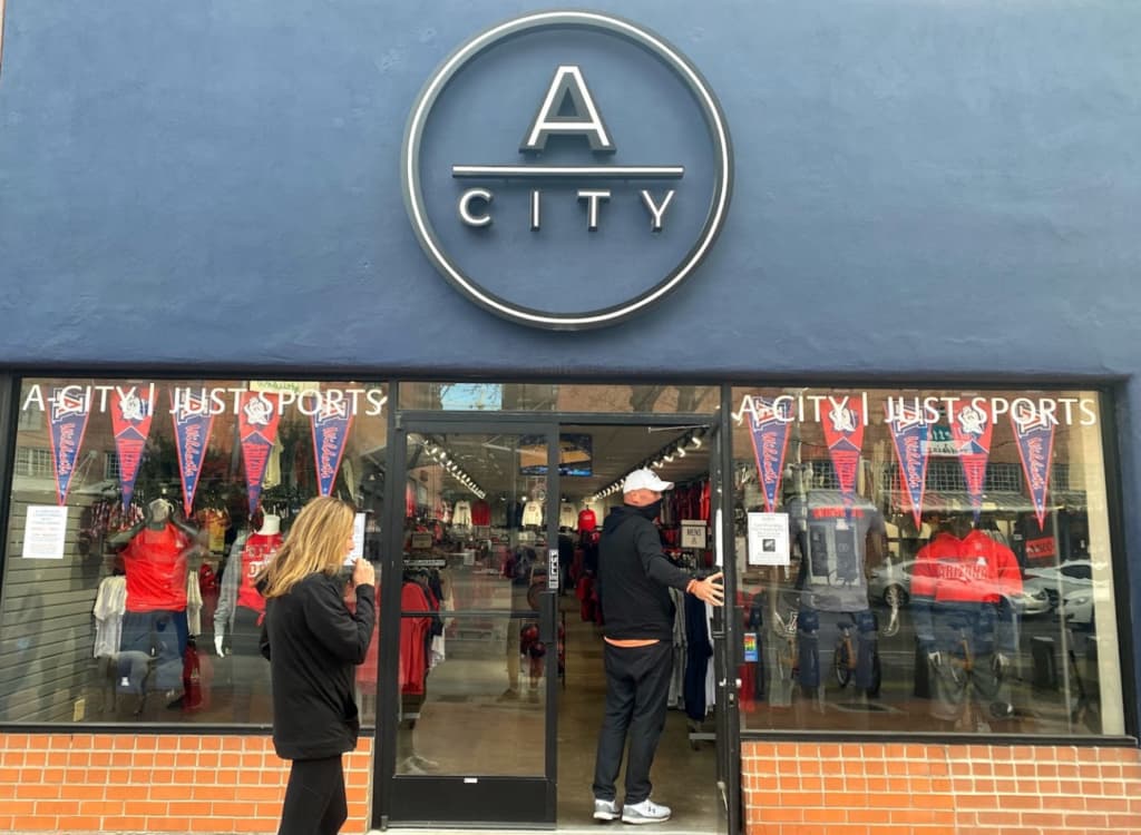 A-City Logo above a door. Person holding the door open for another person. Windows with sports flags hanging in the windows as well as mannequins display attire.