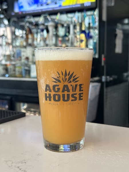 Full beer glass, with the Agave House logo on it. Bar in the blurred background.